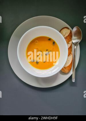 Hausgemachte frische Kürbissuppe garniert mit Kürbiskernen und Leinöl als Topping und Brezel auf einem Teller mit Suppenschüssel und Löffel Stockfoto