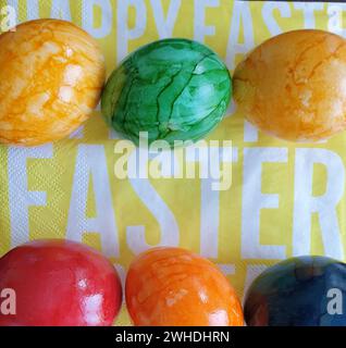 Gekochte bunte Eier in den Eierfarben Gelbgrün Blau und Rot liegen auf einer gelben Serviette mit weißem Schriftzug für Ostern Stockfoto