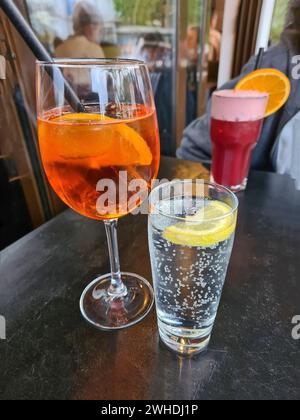 Das alkoholische Erfrischungsgetränk Aperol Spritz steht auf einem Tisch neben einem Glas Mineralwasser und einem alkoholfreien roten Fruchtcocktail im Hintergrund Stockfoto