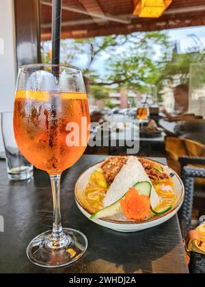 Ein orangenfarbenes Sommergetränk mit Alkohol im Glas steht neben einem asiatischen Gericht im Restaurant Stockfoto