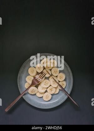 Geschnittene Bananenscheiben auf einem hellblauen Teller mit Messer und Gabel als gesundes Dessert vor dunklem Hintergrund Stockfoto