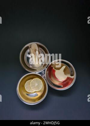 Gebissene Apfelscheibe in Scheiben geschnittene BananenZitronenscheibe in einer der drei silbernen Schalen vor einem grauen Diätthema Stockfoto