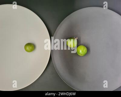 Eine weiße Platte mit 1 grünen Tomaten und eine graue Platte mit 2 kleinen Tomaten auf grauem Hintergrund zum Thema Kontraste und Minimalismus Stockfoto