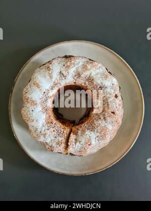 Runder Marmorkuchen mit Puderzucker und einem Loch in der Mitte steht auf einer cremefarbenen Keramikplatte vor dunkelgrauem Hintergrund Stockfoto