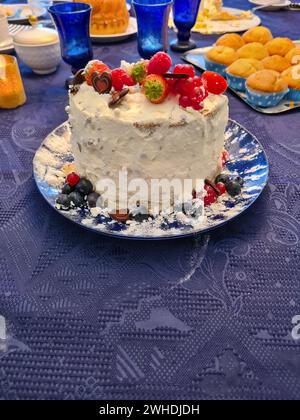 Auf einer blauen Tischdecke steht ein festlicher Sahnekuchen mit Früchten wie Erdbeeren, Himbeeren, Johannisbeeren und Heidelbeeren als Dekoration Stockfoto