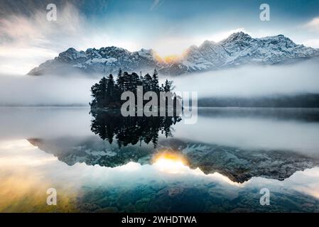 Sonnenaufgang mit Nebelschwaden über dem Eibsee mit Blick auf die Sasseninsel und das Zugspitzmassiv an einem kalten Herbsttag. Zugspitze, Garmisch-Partenkirchen, Bayern, Süddeutschland, Deutschland. Stockfoto