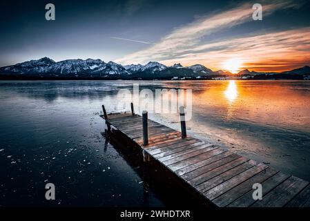 Blick von einem Holzsteg über den gefrorenen, winterlichen Hopfensee im Ostallgäu mit Blick auf die Allgäuer Alpen und den Sonnenuntergang. Ostallgäu, Allgäu, Bayern, Deutschland Stockfoto