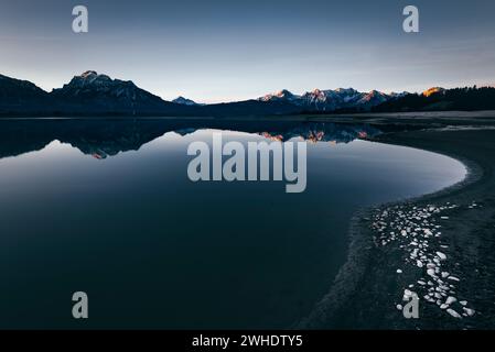 Alpenpanorama mit Reflexion im teilweise trockengelegten, winterlichen Forggensee bei Füssen im Allgäu. Alpenglow im ersten Sonnenlicht. Im Vordergrund, Steine auf dem gefrorenen Seegrund. Stockfoto