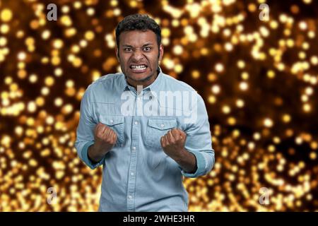 Ein wütender aggressiver Kerl, der grimmig und Fäuste schüttelt. Festlicher Bokeh-Hintergrund. Menschliche negative Ausdrücke. Stockfoto