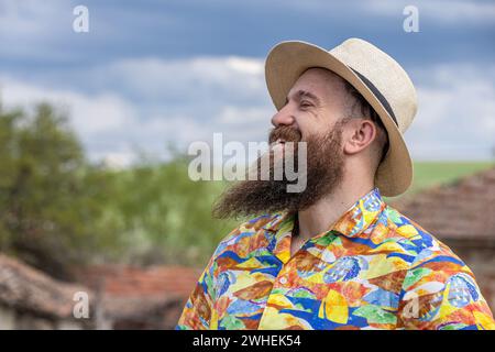 Junger bärtiger Mann mit Fedorahut, der glücklich lächelt. Nahporträt eines bärtigen jungen Mannes, der legere Kleidung trägt und mit einem in die Kamera schaut Stockfoto