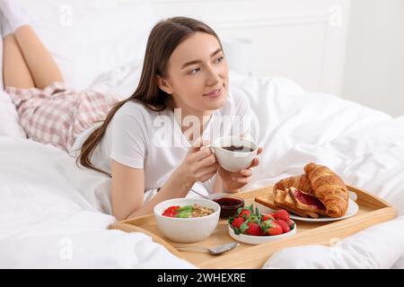 Schöne Frau, die Kaffee in der Nähe des Tabletts mit Frühstück auf dem Bett trinkt Stockfoto