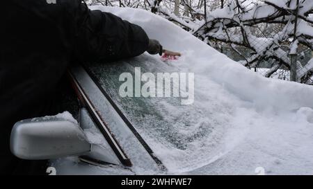 Eine Person in warmer schwarzer Kleidung räumt nach schlechtem Wetter im Winter eine Schneeschicht von der Windschutzscheibe einer grauen Maschine und reinigt das Auto vom Schnee Stockfoto