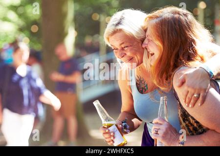 Lachend, reif oder befreundet mit Bier beim Musikfestival für ein Konzert oder eine Party im lustigen Urlaub zusammen. Lächeln, Frauen oder glückliche Menschen, die Drinks genießen Stockfoto