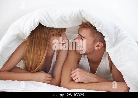 Glückliches Paar, romantisch und Moment im Bett, zusammen und aufgeregt auf die Ehe, glücklich und entspannt. Partner, sprechen und lächeln mit Liebe, Heim und Lügen Stockfoto