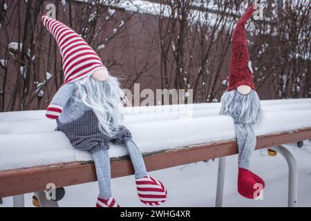 Ein paar Zwerge auf der Bank im Schnee. Romantisches Geschenk. Freundschaftskonzept. Valentinstag-Konzept. Gnome mit Herz und rotem Hut im Schnee. Stockfoto