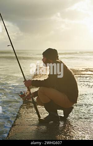 Mann, Warten und Angeln am Strand in der Natur, Entspannung und natürliche Meer Hobby für Wellness im Urlaub. Fischer, reisen oder Urlaub in kapstadt auf dem Meer Stockfoto