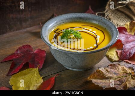 Herbstsuppe aus rotem kuri-Kürbis mit Petersilie-Garnitur in einer rustikalen Schüssel auf einem dunklen Holztisch mit bunten Blättern für Thanksgiv Stockfoto