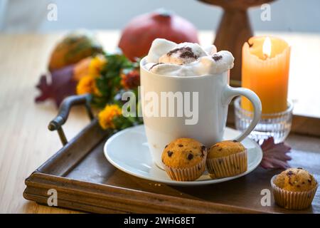 Weißer Becher mit heißem Schokoladengetränk und schmelzenden Marshmallows, Muffins, Kerze und Herbstdekoration auf einem Tisch, Kopierraum, Auswahl Stockfoto