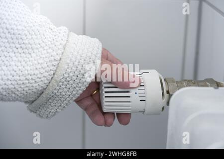 Die Hand einer Frau in warmer Kleidung reduziert die Wärme am Thermostat des Heizkörpers aufgrund der Energiekrise mit steigenden Preisen. Stockfoto