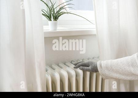 Eine Frau mit warmer Kleidung und Handschuhen legt ihre Hand auf eine alte Heizung, um die Temperatur zu spüren, und spart Energie durch Aufblasen und Ri Stockfoto