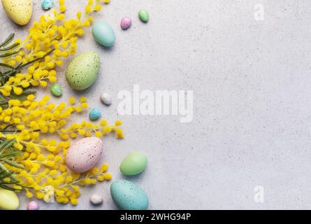 Dieses Bild zeigt ein festliches Osterarrangement aus pastellfarbenen Eiern und hellgelben Mimosa-Blüten auf einer strukturierten hellgrauen Oberfläche. Stockfoto