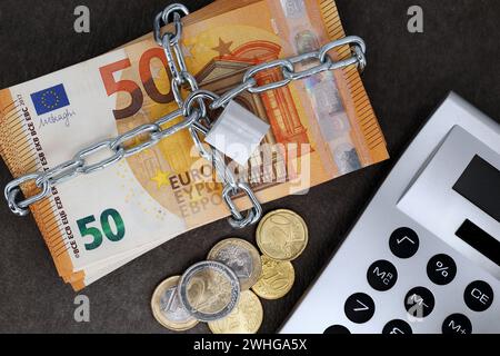 Ein Bargeldstapel mit 50-Euro-Banknoten ist mit einer Kette und einem Vorhängeschloss neben einem Taschenrechner und Münzen verschlossen Stockfoto