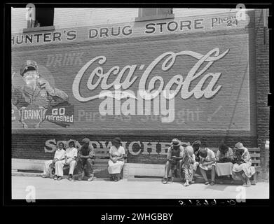 Allgemeine Szene, Hauptstraße. Greensboro, Georgia, USA, um 1930er Jahre Die Leute sitzen auf der Straßenbank in der Nachmittagssonne unter einer großen Coca-Cola-Werbung, die auf der Ziegelwand von Hunter's Drug Store gemalt ist Stockfoto