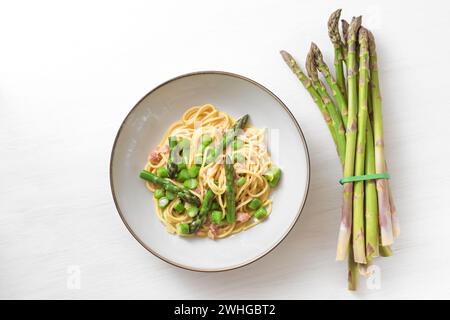 Grüner Spargel mit Spaghetti, Speck und Carbonarasauce auf einem Teller neben einem Bündel rohen Gemüses auf einem weiß bemalten Woo Stockfoto