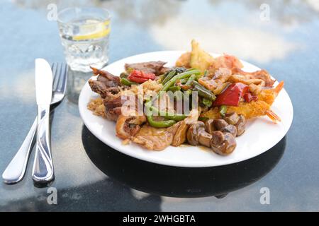 Chinesisches Gericht in einem Straßenrestaurant, verschiedene Fleisch- und Gemüsesorten mit gebratenem Reis auf einem weißen Teller, Besteck und ein Glas mit Wasser Stockfoto