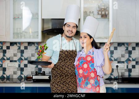 Glückliches indisches Paar in Kochuniform mit Schürze, Kochmütze bereit, leckeres Essen zuzubereiten. In der Küche mit Pfanne, Pin bela Stockfoto