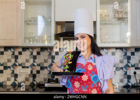 Wunderschöne indianerin in Kochuniform mit Schürze, Kochmütze bereit, leckeres Essen zu machen. In der Küche stehend mit Antihaftpfanne mit Fliegen Stockfoto