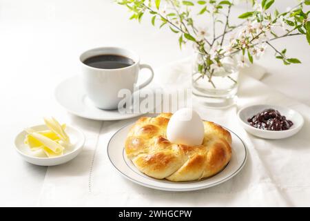 Osterhefe in Nestform mit Ei, Kaffee, Butter und Marmelade als festliches Sonntagsfrühstück auf einem weißen Tisch, Kopie Stockfoto