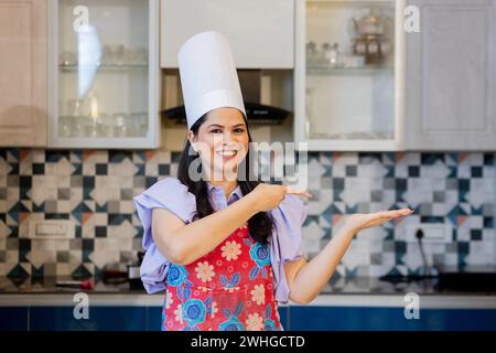 Wunderschöne indianerin in Kochuniform mit Schürze, Kochmütze in der Küche mit einer Hand ausgestreckt und die andere Hand zeigt auf die Stockfoto