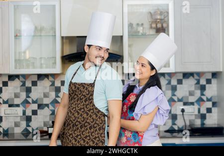 Glückliches indisches Paar in Kochuniform mit Schürze, Kochmütze bereit, leckeres Essen zuzubereiten. In der Küche stehen und Spaß haben. Stockfoto