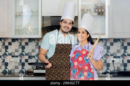 Glückliches indisches Paar in Kochuniform mit Schürze, Kochmütze bereit, leckeres Essen zuzubereiten. Steht in der Küche mit Pfanne, rollt Pin belan in ha Stockfoto