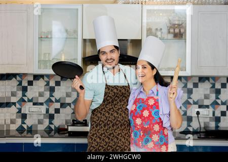 Glückliches indisches Paar in Kochuniform mit Schürze, Kochmütze bereit, leckeres Essen zuzubereiten. Steht in der Küche mit Pfanne, rollt Pin belan in ha Stockfoto