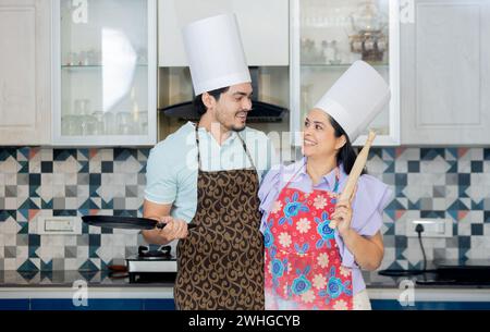 Glückliches indisches Paar in Kochuniform mit Schürze, Kochmütze bereit, leckeres Essen zuzubereiten. Steht in der Küche mit Pfanne, rollt Pin belan in ha Stockfoto