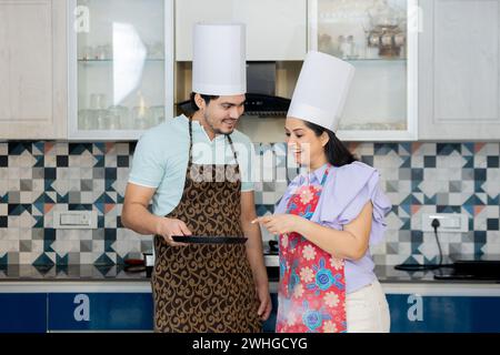 Glückliches indisches Paar in Kochuniform mit Schürze, Kochmütze bereit, leckeres Essen zuzubereiten. In der Küche mit Pfanne in der Hand stehen. Kochen Wird Angezeigt Stockfoto