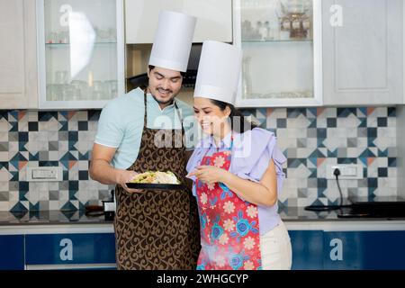 Glückliches indisches Paar in Kochuniform mit Schürze, das leckeres Essen in der Küche zubereitete. Kochfähigkeiten Zeigen Stockfoto