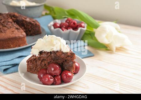 Saftiger hausgemachter Schokoladenkuchen mit morello-Kirschen und Schlagsahne, einigen Tulpenblüten und einer blauen Serviette auf einem hellen hölzernen t Stockfoto