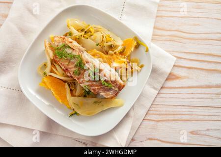 Kabeljau-Filet auf Fenchelgemüse mit Orangenscheiben und Fenchelgrüner Garnitur auf weißem Teller und Serviette, heller Holztisch Stockfoto