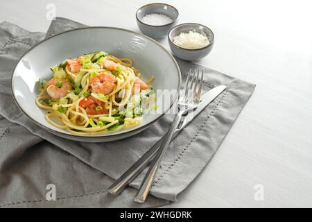 Spaghetti mit Zucchini-Zucchini, Frühlingszwiebeln und Garnelen auf einem grauen Teller auf einem weißen Tisch mit Serviette und Besteck, gesund Stockfoto