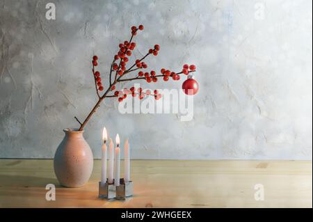 Minimalistischer zweiter Advent mit vier kleinen Kerzen, zwei brennend und ein Beerenzweig mit roter weihnachtskugel in einer Vase, verschneit Stockfoto