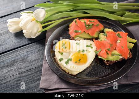 Romantische Mahlzeit mit Liebe, Spiegeleier in Herzform, Sandwiches mit Avocado und geräuchertem Lachs, weiße Tulpen auf einem dunkelblauen Rusti Stockfoto