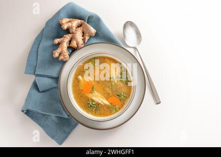 Gesunde Suppe aus Hühnerfleisch, Gemüse und Ingwer in einer Schüssel auf einer blauen Serviette und einem schwulen weißen Hintergrund, von oben aus Stockfoto