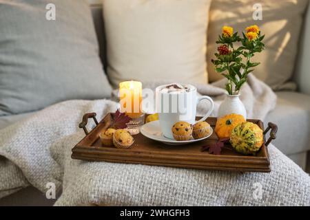 Heißes Schokoladengetränk mit Marshmallows und Mini-Muffins auf einem Tablett mit Kerze, Blumen und Herbstdekoration neben einer gemütlichen Couch Stockfoto