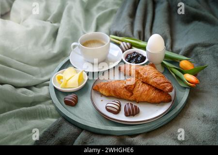 Frühstück mit Liebe im Bett, Kaffee, Croissant, Marmelade, Ei, ein paar Schokoladenherzen und Tulpen auf einem Tablett auf graugrünem Bett lin Stockfoto