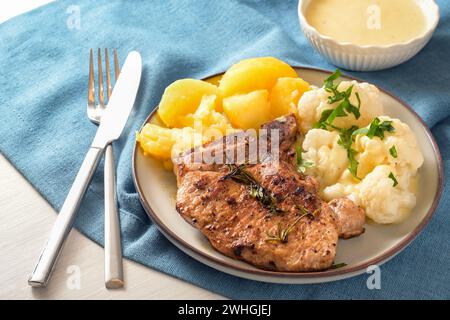 Gericht mit Schweinekotelett, Blumenkohl, Kartoffeln und Hollandaise-Sauce serviert mit Kräutergarnitur auf einem Teller und einer blauen Serviette, ausgewählt Stockfoto