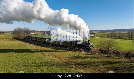 Ein Blick auf die Drohne eines antiken Dampfeisenzugs, der sich durch die Landschaft und das Ackerland nähert und Rauch auf einem sonnigen Wi bläst Stockfoto