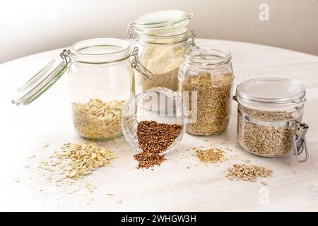 Getreide und Samen wie Haferflocken, Leinsamen, Sesam und Sonnenblumenkerne in Glasgläsern auf einem hellen Holztisch, Gesundheit Stockfoto